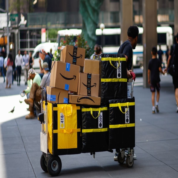 An Amazon delivery person pulling a large amount of packages on a wheeled cart on New York City sidewalk.