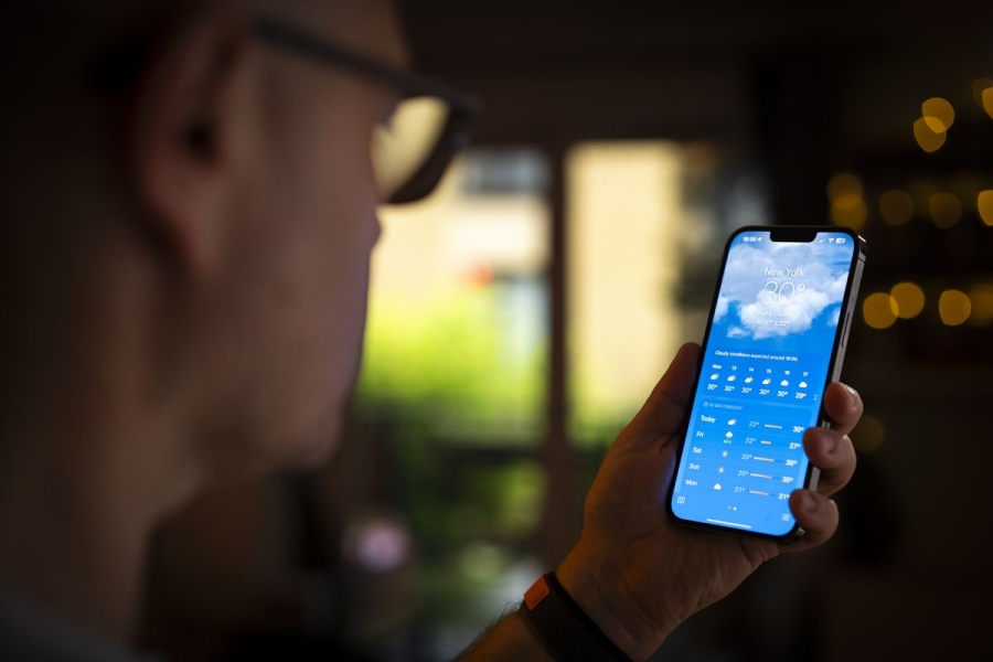 A man wearing glasses looks at the phone in his hand, which shows Apple’s weather app on the screen.