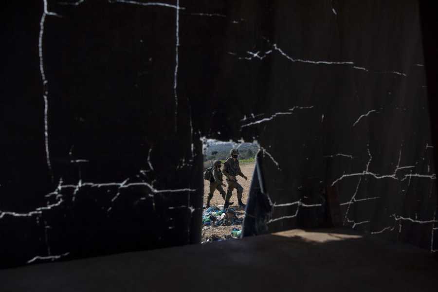 Seen through a small hole in the wall, soldiers in camouflage military uniforms and helmets walk past. 