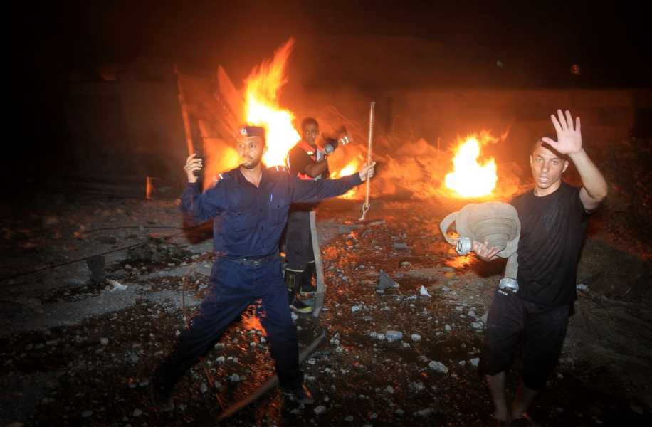 Fires swirl, illuminating the night, as a fireman holds a hose, looking over his shoulder to as if communicating with someone outside of the frame. A blue uniformed policeman gestures toward the flames as a man in all black holds a length of hose.