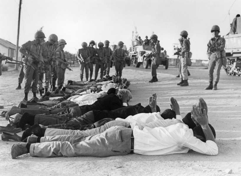 On a desert plain, a group of Israeli soldiers in fatigues and bucket helmets point rifles at men in slacks and shirts who are lined up on the ground, their hands clasped atop their heads. Tanks and troop transports roll along in the background.