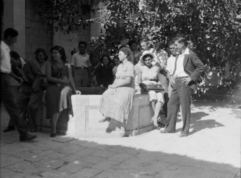 Young Jewish students, in suits and dresses, sit in a courtyard. The sun shines on a group of students hanging around a fountain; ivy clings to a wall behind them. A few students sit in the shadows. Many appear to be engaged in conversation.