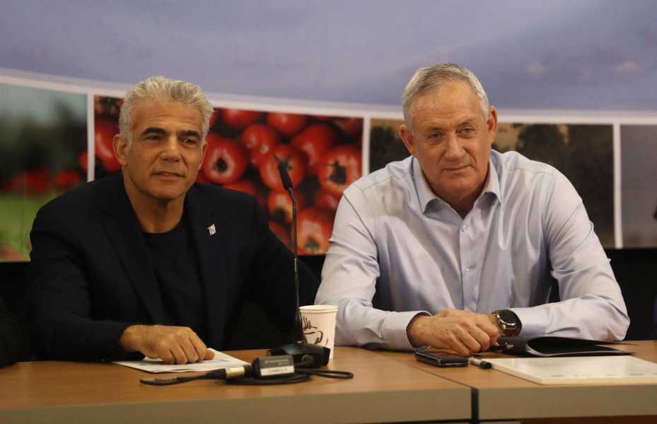 Gantz, in a light blue collared shirt, and Lapid, in a black shirt and blazer, both smile and listen, sitting at a large wooden table.