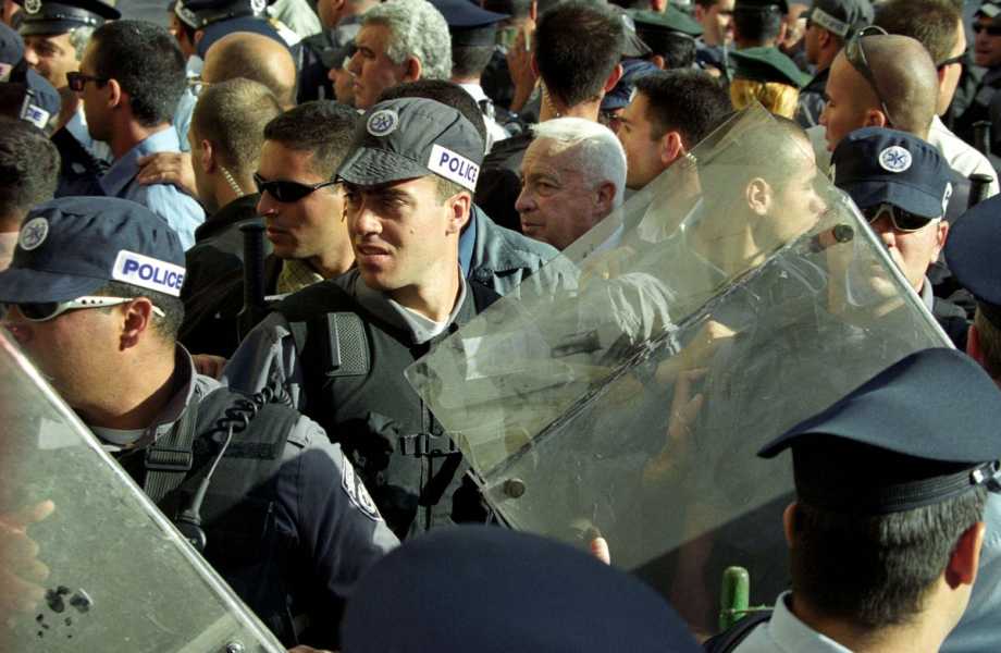 Sharon, white haired, can barely be seen, ensconced in a tangle of police with armored vest and riot shields. Together, they proceed in a tight knot in golden light.