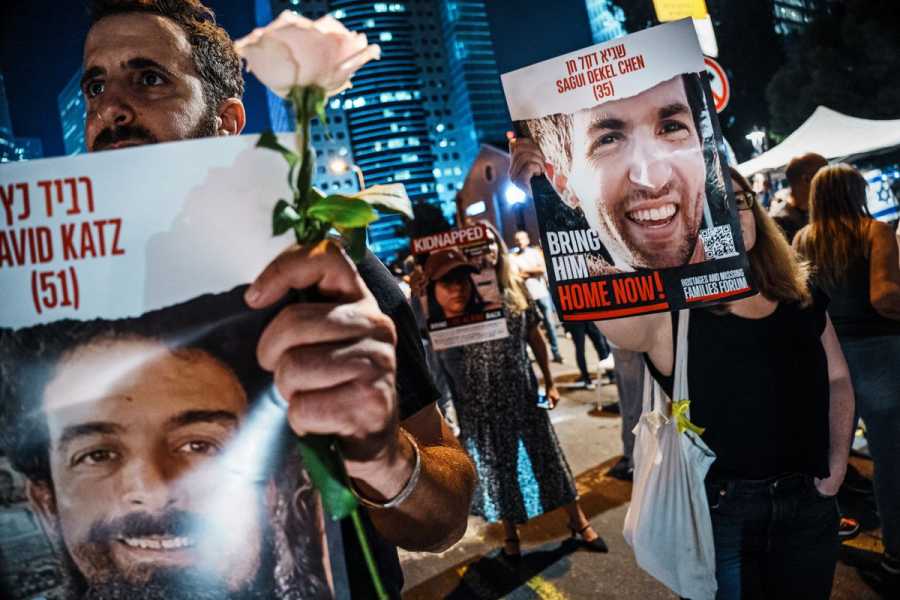 At night, men and women dressed in black hold posters with color photos of various hostages, each bearing the name of the person in the photo in English and Hebrew, their age, and the words “Bring Him Home Now!”