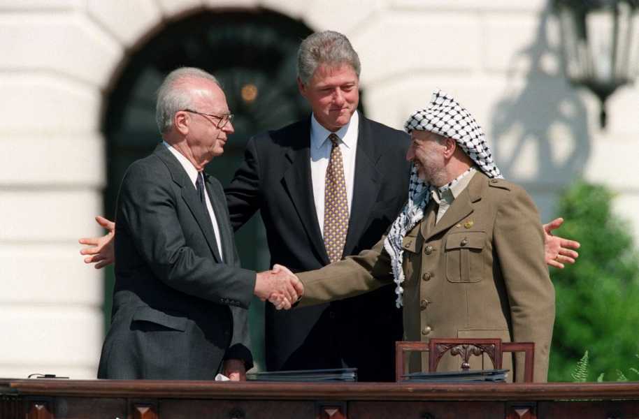 Then US President Bill Clinton, gray haired and clean shaven in a dark suit and black and gold tie, stands behind then Israeli Prime Minister Yitzhak Rabin (white haired in a dark suit) and then PLO leader Yasser Arafat&nbsp;(in green fatigues and a black and white keffiyeh) as the two Middle Eastern leaders shake hands.