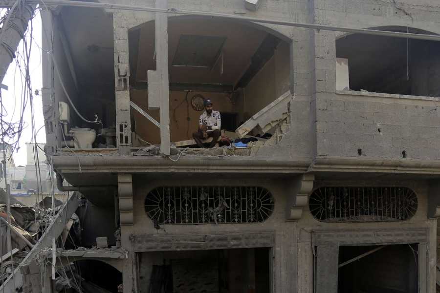 A man sits on the second floor of a partially destroyed building, which is missing a wall. The missing wall exposes the building’s interiors, including a toilet and sink.