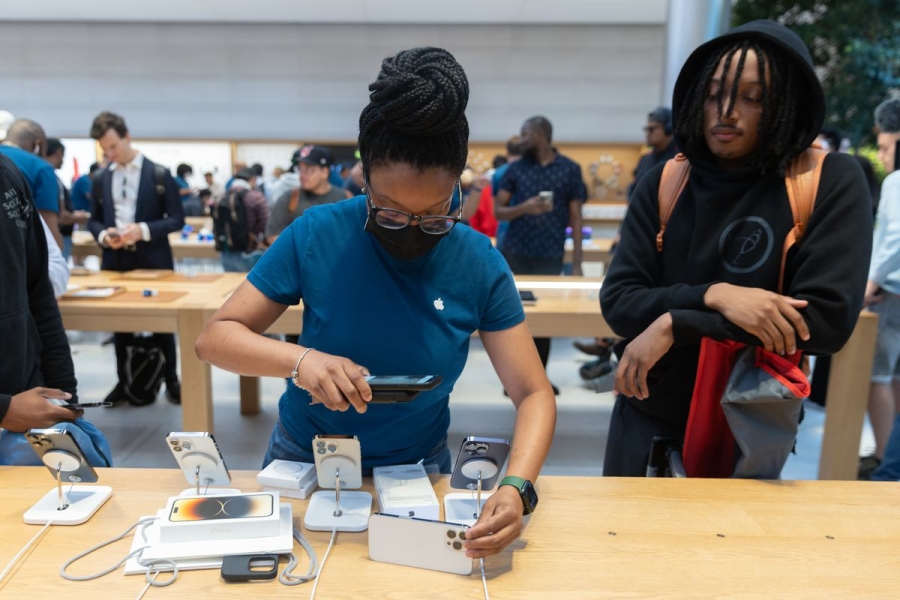 An Apple Store employee scans an iPhone with a handheld reader while a customer stands and watches.