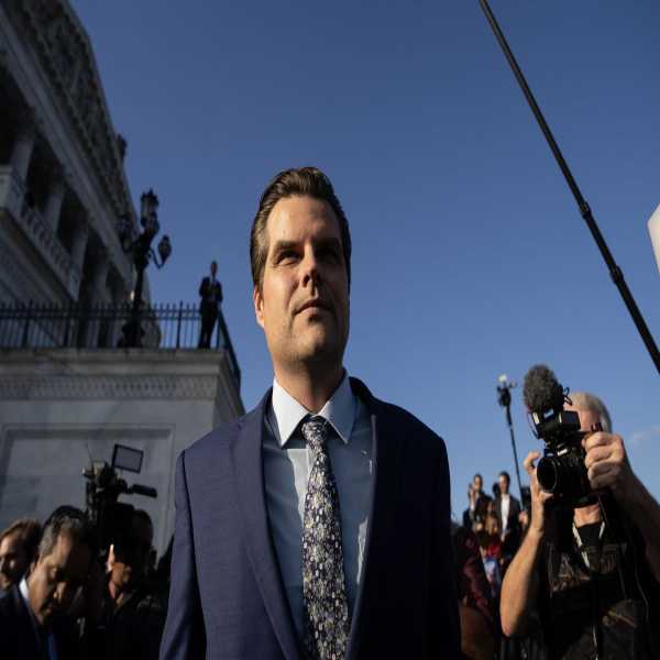 Seen from below, Gaetz, clean shaven with his dark hair slicked back, smiles slightly. The setting sun illuminates half of his face, leaving the other half in shadow. Reporters with their recorders, phones, and cameras cluster around him. Behind them, in shadow, is the Capitol building.