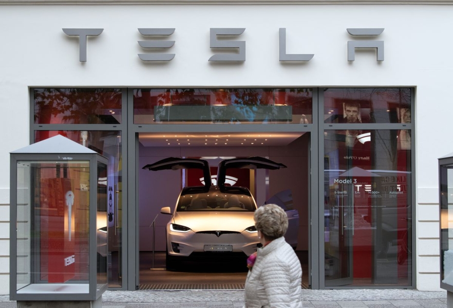 A pedestrian walks past a Tesla showroom displaying a Tesla Model 3 with its gull-wing doors open.