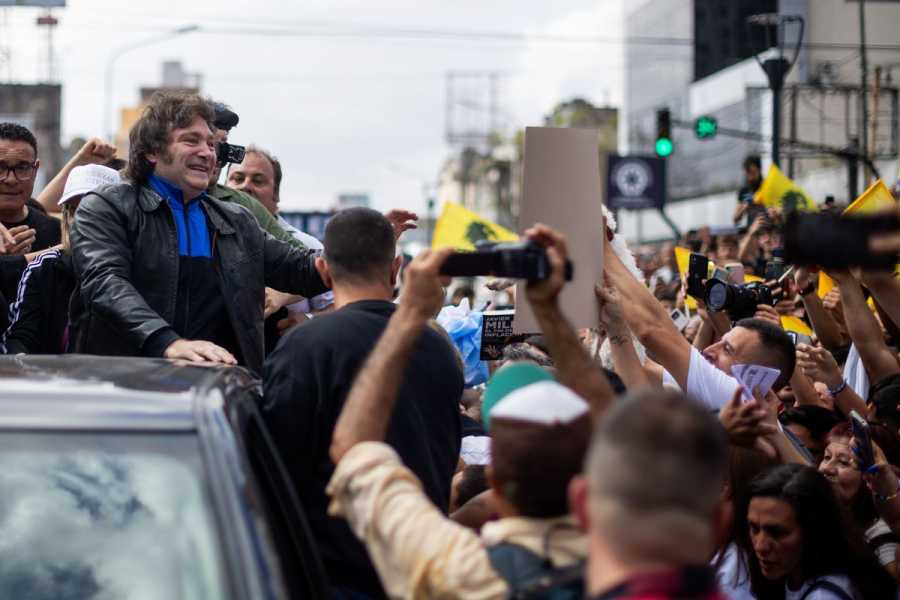 Milei stands in the bed of a truck, waving and smiling at a crowd, many of whom wave small yellow flags and take photos. 