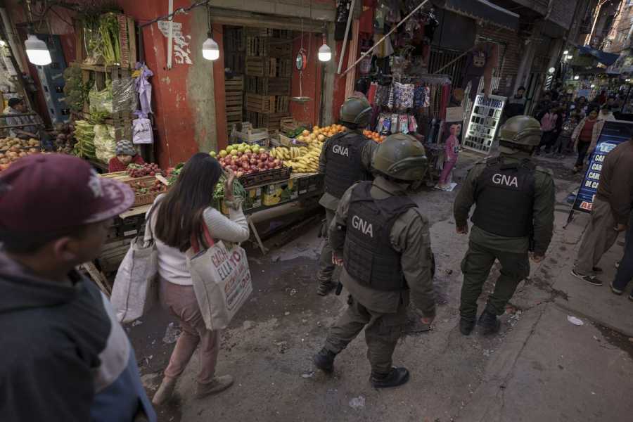 National Gendarmerie guards walk past market stalls full of fruits and vegetables, wearing army green uniforms, helmets, and black flak jackets.