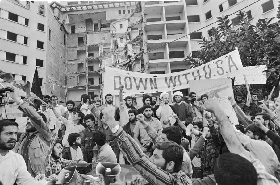 In a black-and-white photo, a crowd of young men raise their fists and cheer; a few hold a large banner that reads: “Down with U.S.A,” while others hold a poster featuring the Hezbollah logo, the group’s name, with one of the letters raising up above the word to form a fist grasping a rifle. In the background, the crumbling US embassy is visible.