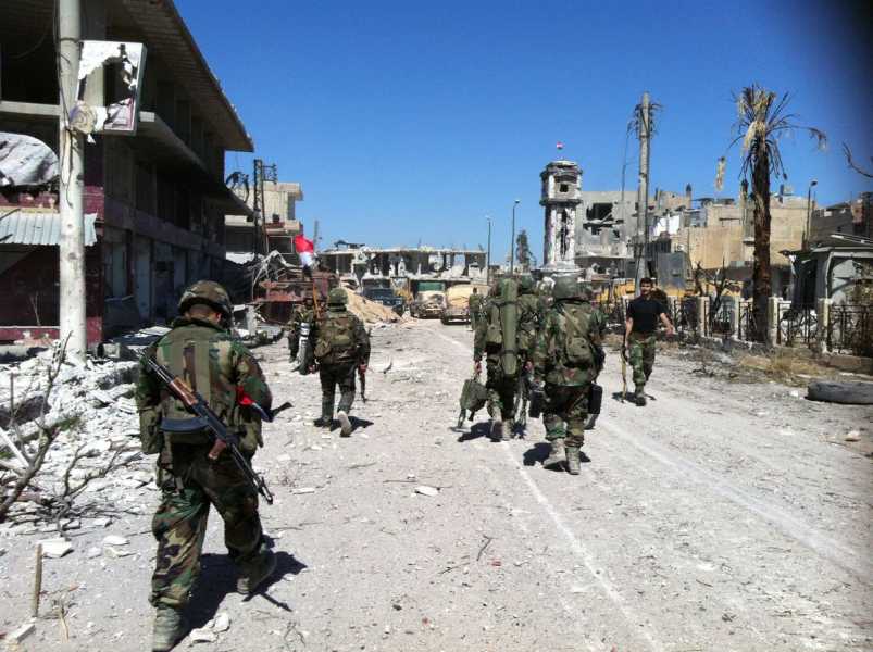 Soliders in camouflage and bucket helmets, laden with weapons and large bags on their backs, walk down an empty street, electric blue sky above them. Buildings and trees still stand around them, though the structures have lost all their walls and windows, and the trees many of their leaves in the intense fighting.