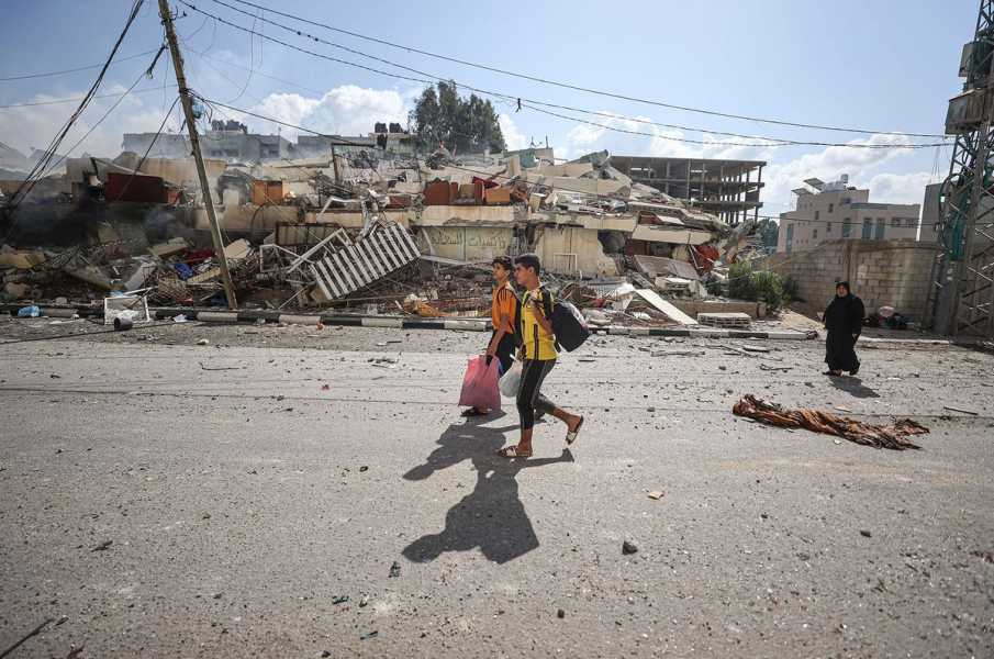 Two young men and a woman walk down an asphalt road, past the piled rubble of collapsed houses. 