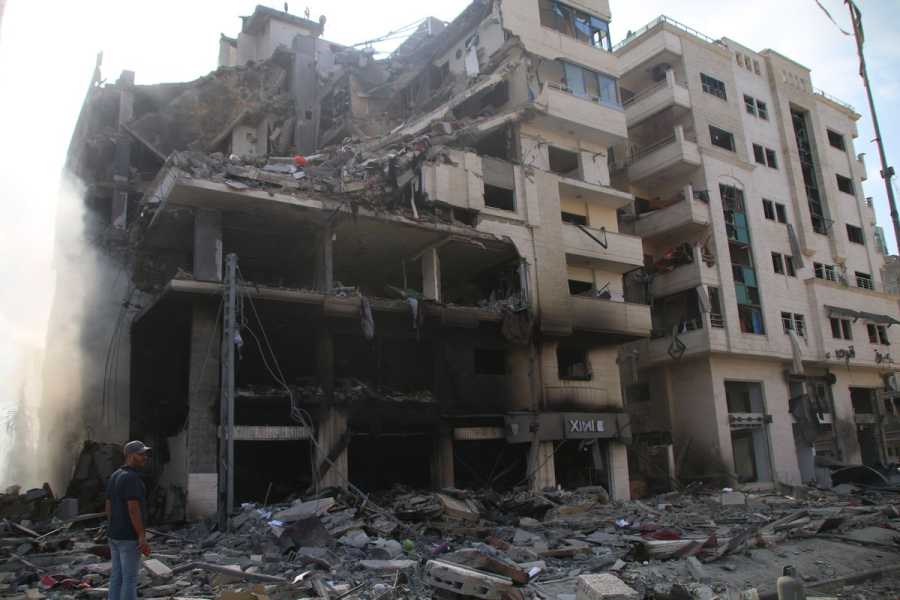 A man stands in the left side of the frame in front of a building that has a crater in the upper left side of it. There is rubble everywhere.