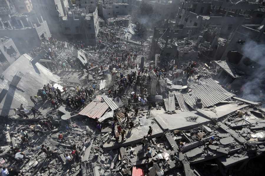 Seen from above, some buildings have been flattened to the ground, while a few still stand, missing windows, walls, and roofs. Crowds stand among the gray rubble, concrete slabs, and sheets of metal. 