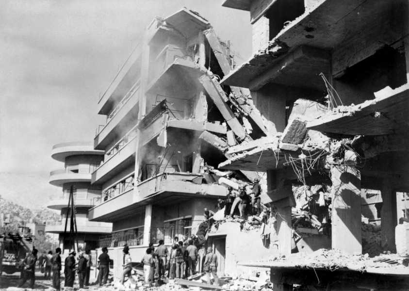 An apartment building looms above a crowd, its left and right sides intact, its middle sunken under the weight of an Israeli attack.