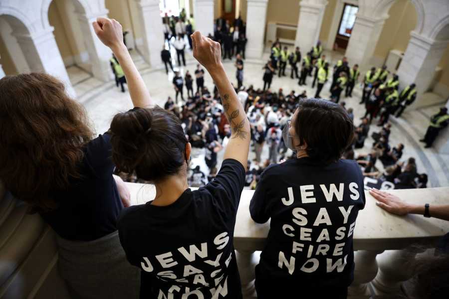 Demonstrators look down on a crowd below from the rotunda balcony, lifting their fists in the air. They wear black t-shirts printed with the words  “Jews Say Ceasefire Now” in white capital letters.