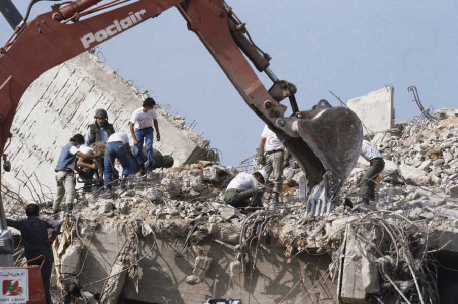 The arm of a dusty red digger stretches across the frame as men in Red Crescent T-shirts and marines half in their uniforms, half in civilian clothes sort through a massive pile of concrete and metal. They stand on what seems to be the second or third floor of a very unstable building, roofless and without walls, blue sky visible above them.
