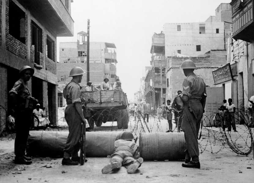 On a civilian street, soldiers in fatigues and bucket helmets are arranged behind a barbed wire and barrel barricade, a military vehicle carrying more troops parked just beyond the checkpoint. Egyptians mill about, some beneath a store sign written in Arabic.