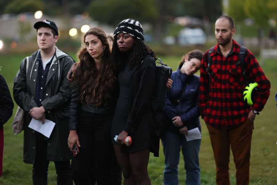 A group of people stand outside on a grassy lawn, wearing somber expressions. Two women stand with their arms around each other. Behind them, a woman leans her head against a man’s shoulder. 