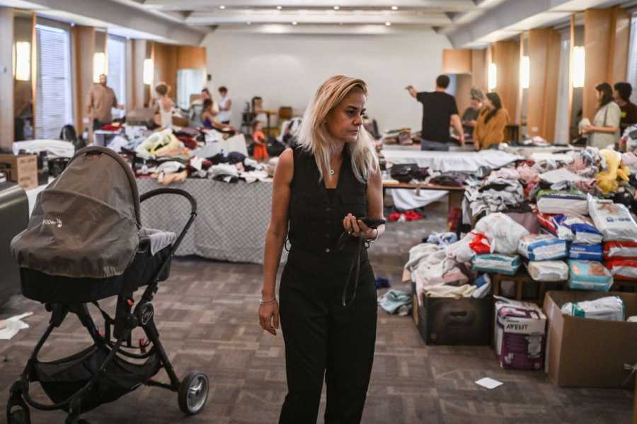 People look through tables of clothing of all kinds, piles of baby equipment and diapers, while volunteers direct movement in the background. A woman in a black jumpsuit with a phone in her hand walks past the tables in the foreground.