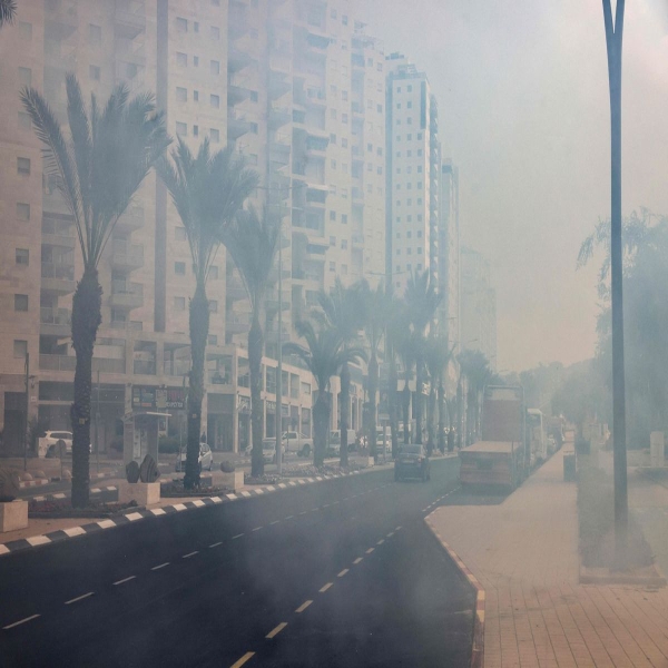 A photograph of a palm-tree-lined street in Ashkelon, in southern Israel. The light is dimmed by smoke from a nearby fire.