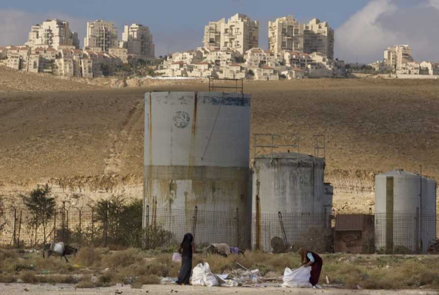 Women surrounded by dry, barren brown land, fill white trash bags. Behind them stand some industrial silos, and farther in the distance, a settlement of multistory buildings can be seen. 