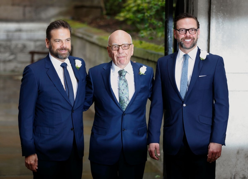 Rupert Murdoch stands between his sons Lachlan and James at a wedding ceremony. All are wearing blue suits, ties, white shirts, and white rose boutonnieres. 