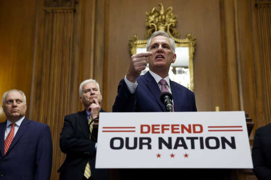 McCarthy speaking at a podium with a sign that says “defend our nation.”