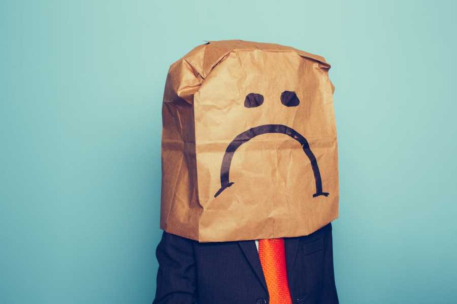 A man in a suit wears a paper bag on his head with a frowning face painted on.