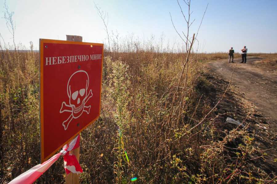 A red sign with a skull and crossbones reads, in Russian, “Danger. Mines.” It appears on a fence in front of an open field beside a dirt road.