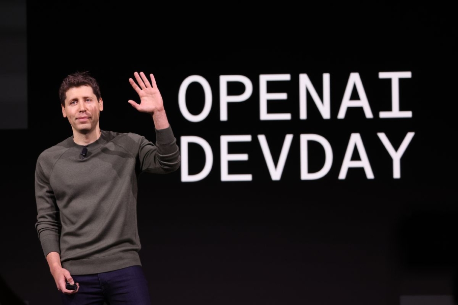 Sam Altman waving from onstage at OpenAI’s DevDay.