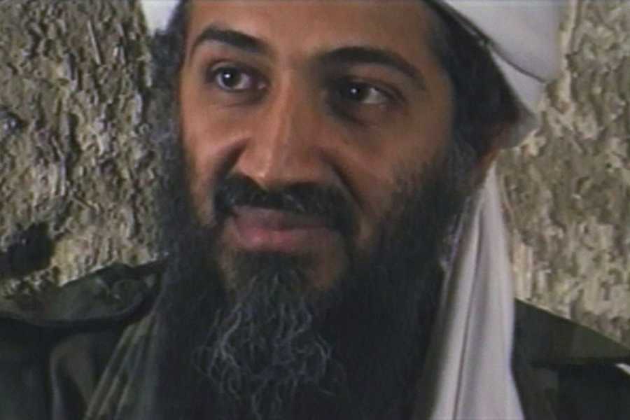 A close-up of bin Laden’s face; it is long, thin. His skin is dark brown, and he sports a thick salt-and-pepper beard as well as a white turban. He appears to be wearing combat fatigues.
