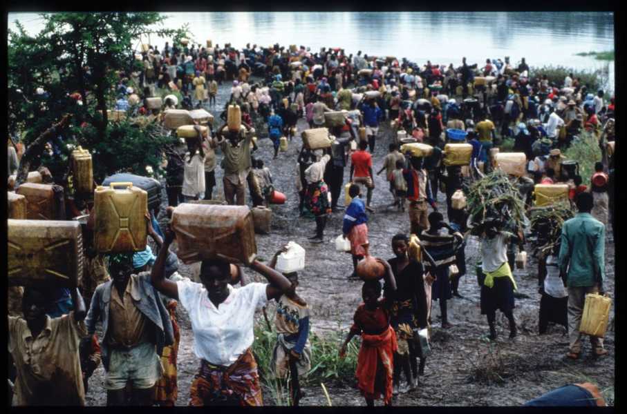 Crowds of people gather on a muddy riverbank, most holding belongings or balancing them on their heads.