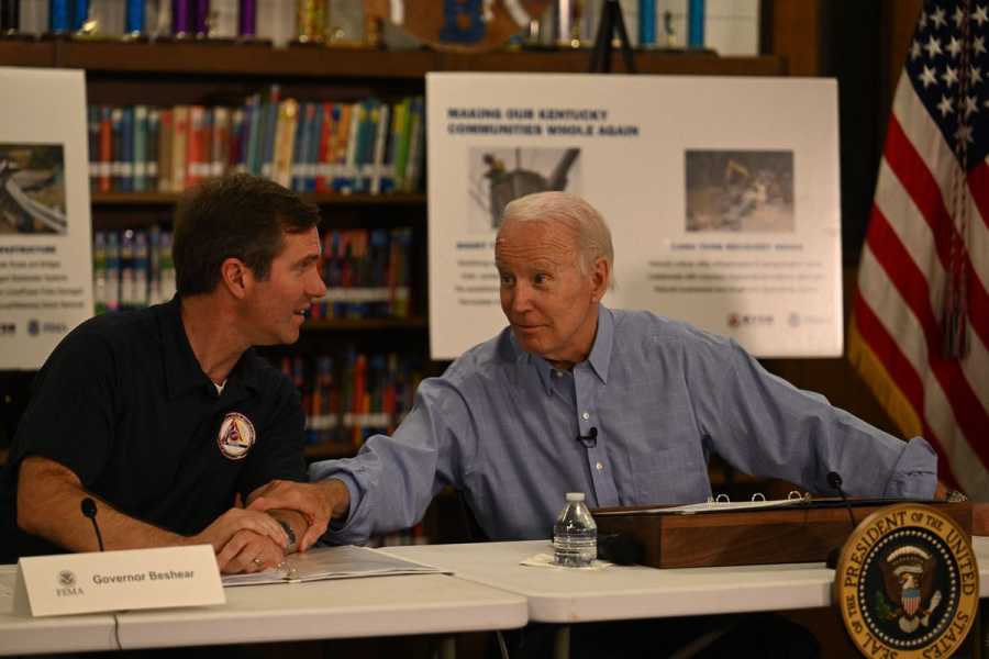 Beshear and Biden sit at a table, and Biden is leaning over to grab Beshear’s arm while they talk.