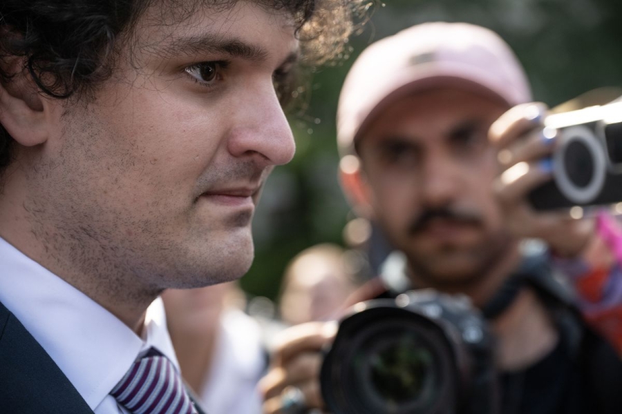 Sam Bankman-Fried, up close in profile, with dark curly hair and a black suit, looks toward reporters with cameras and a crowd of people.