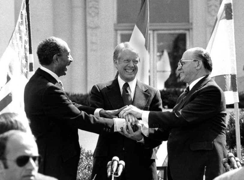 In a black-and-white photo, the three men, all dressed in dark suits, clasp hands while smiling broadly. The flags of their respective nations wave behind them.