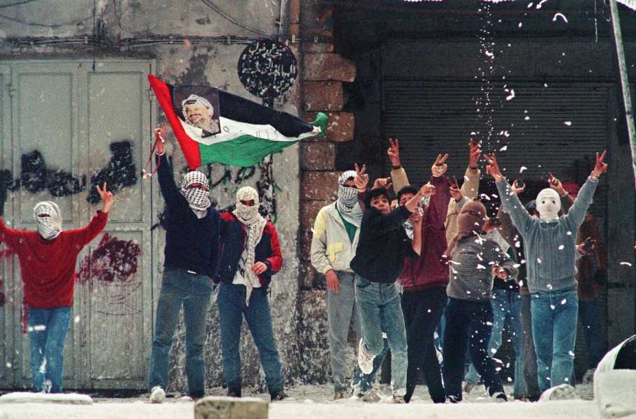 What appear to be a gaggle of teen and pre-teen boys, some with their faces covered by keffiyehs, throw up peace signs on a city street as the white foam falls like snow. One youth waves a Palestinian flag that has a color photo of Yasser Arafat attached to it.