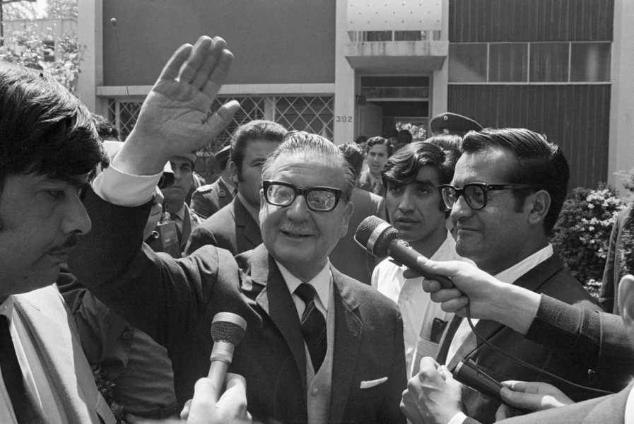 A black-and-white photo shows Chilean President Salvador Allende surrounded by people and waving to the camera.