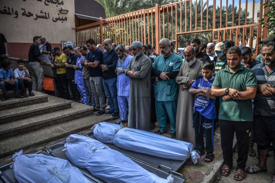 A group of people, including children, stand with heads bowed and arms crossed across their chests in prayer. Before them on the ground lie several body bags. 