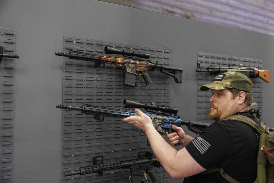 A man in a camouflage hat holds a rifle. More rifles hanging on racks are seen in the background. 