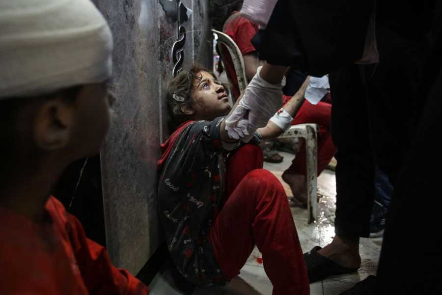 A grade school-aged girl, dressed in red, sits on the floor, her back against the wall. Her face is smeared with dirt. A medical worker stands over her; other patients can be seen both in the fore and backgrounds of the image, waiting to be seen as well.