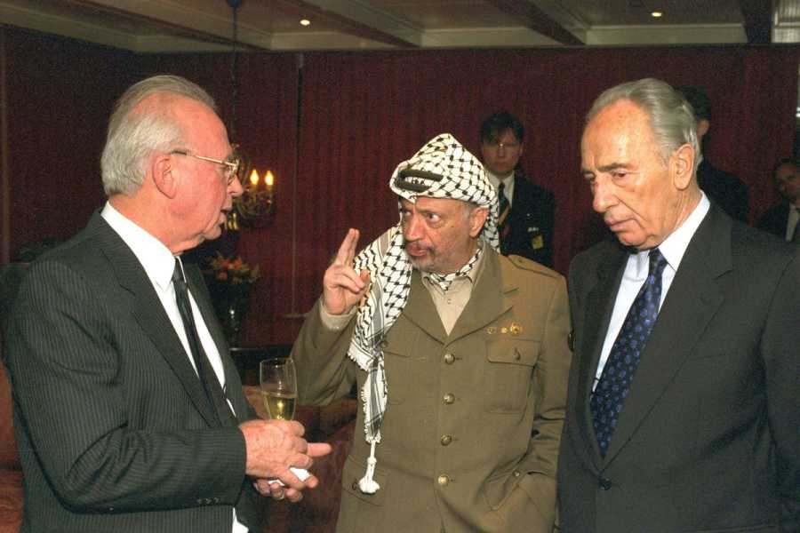 Arafat, in his military uniform and keffiyeh, stands between the Israeli men and holds up two fingers, looking serious. Rabin, in a black suit and tie, holds a glass of champagne, while Peres seems to have his hands in his pockets. Both men listen intently.