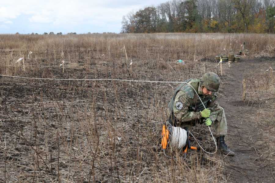 A person in camouflage kneels in a wintry field holding a spool of wire.