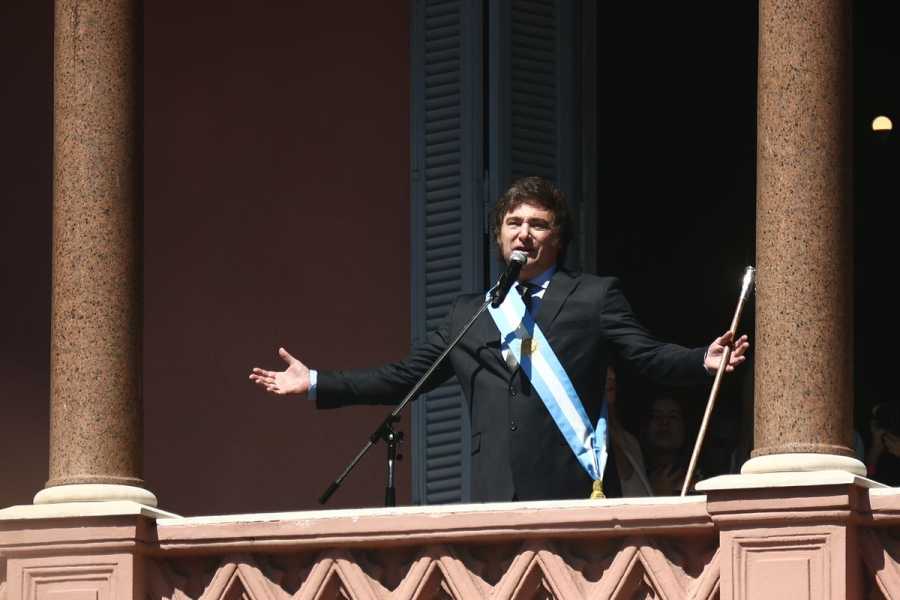 Javier Milei stands on a balcony made of pinkish stone, flanked by two brown marble columns. He raises both hands, holds a scepter, and speaks into a microphone.