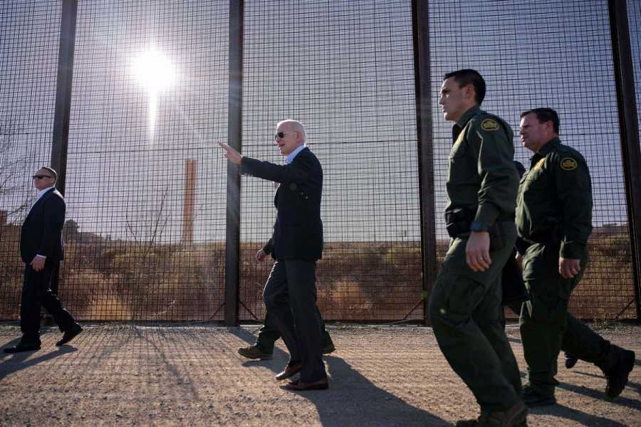 President Biden and several men in uniforms walk past a very tall fence with netting between the posts. 