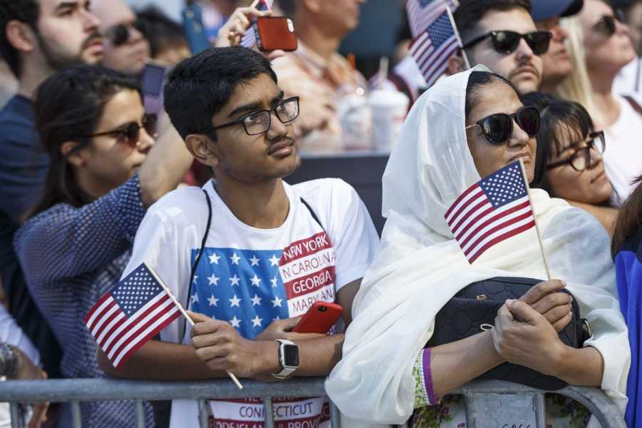 A young man wearing eyeglasses and a T-shirt with an American flag design holds a small American flag in his hand. Beside him, a woman in a white headscarf and sunglasses also holds a small American flag. 