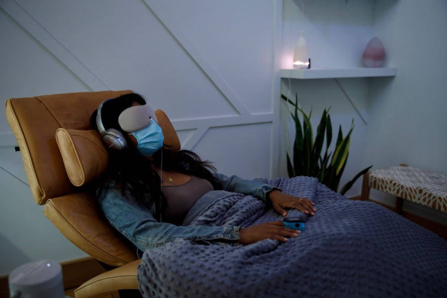 A woman rests on a reclining chair. She wears headphones and an eye mask and has a blanket over her lap. The room is dimly lit.